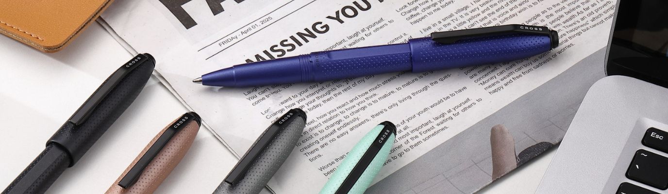 Collection of pens on a newspaper with a laptop in the background